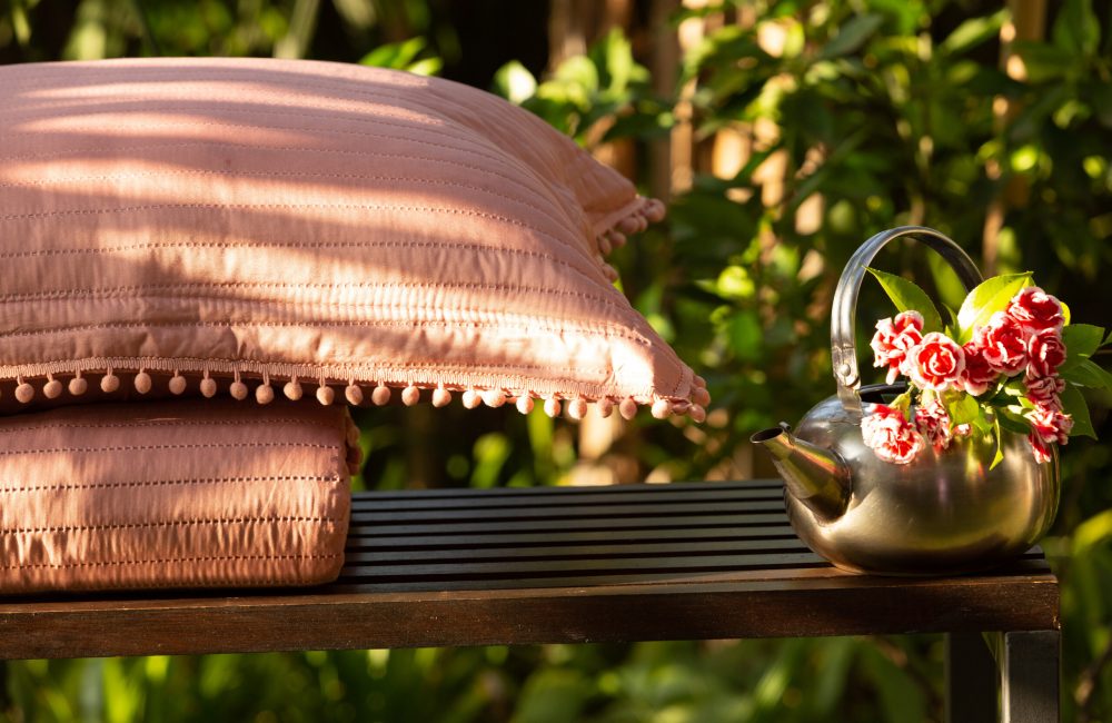 Imagem da Colcha Pompom na cor rosé dobrada em cima de um balcão de madeira escura, com um vaso de flores ao lado.