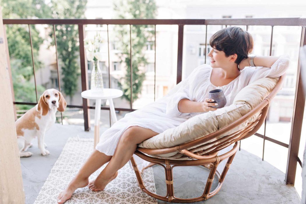 relaxed-barefooted-girl-white-dress-sitting-chair-balcony-holding-cup-tea