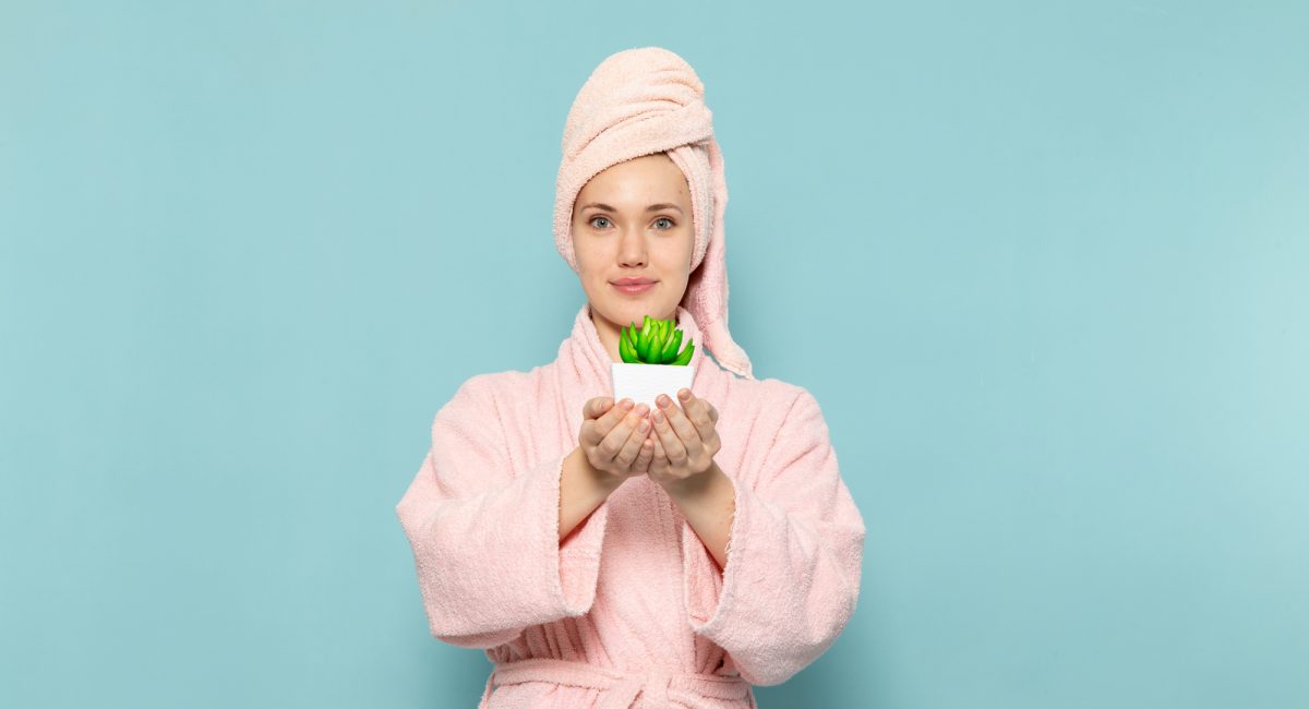 young-female-in-pink-bathrobe-after-shower-holding-little-green-plant-on-blue